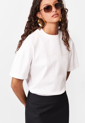 Kobieta o kręconych włosach, nosząca duże złote kolczyki, prostokątne okulary przeciwsłoneczne, biały haftowany t-shirt z napisem "La Monica" oraz czarną spódnicę.