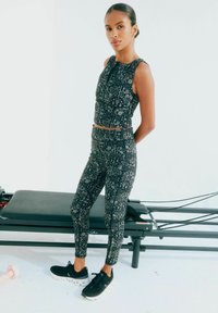 Top corto nero e leggings con motivi bianchi intricati, caratterizzati da un design aderente. Abbinati a scarpe da ginnastica nere. Lo sfondo è bianco semplice.