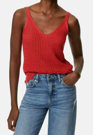 Persona de piel oscura que lleva un top de punto sin mangas rojo con cuello en V y vaqueros azules de cintura alta, con una mano en el bolsillo.