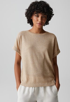 Donna con capelli corti e ricci che indossa una maglia beige a maniche corte e pantaloni bianchi, in piedi con le mani nelle tasche su uno sfondo grigio.