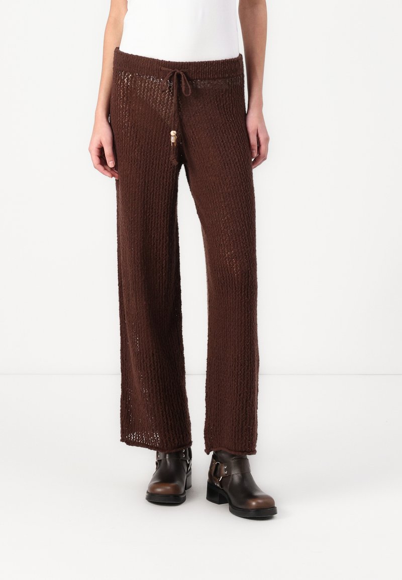 Pantalones de pierna ancha marrones de punto con un patrón texturizado, que cuentan con una cintura ajustable con cordón y detalle de borlas. Combinados con botas negras hasta el tobillo.