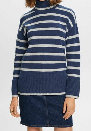 Femme portant un pull à col roulé rayé bleu et gris avec de larges poignets côtelés et une jupe en jean foncé, bras détendus le long du corps.
