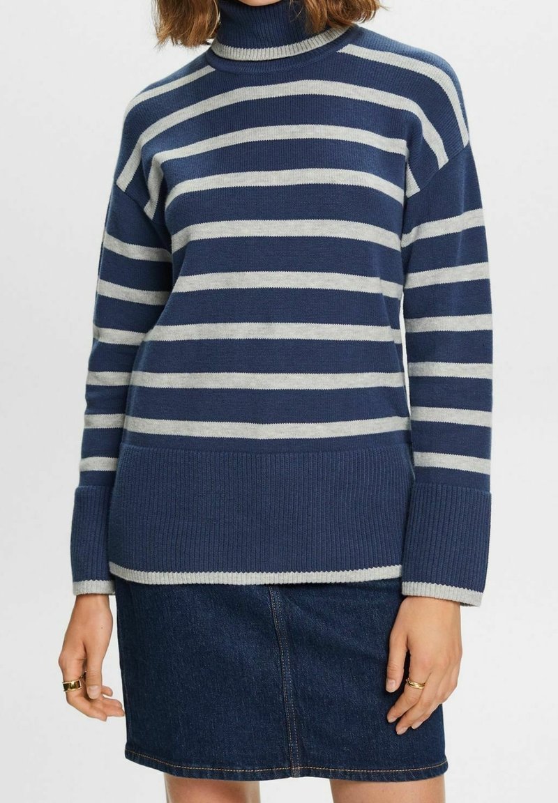 Femme portant un pull à col roulé rayé bleu et gris avec de larges poignets côtelés et une jupe en jean foncé, bras détendus le long du corps.