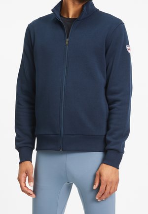 Man in een marineblauwe zip-up jas met een hoge kraag en lichtblauwe strakke sportbroek tegen een witte achtergrond.