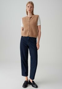 Tanskleurige wolblend sweatervest met een ronde hals en knoopsluiting. Gecombineerd met donkerblauwe ontspannen pasvorm jeans en zwarte instappers.
