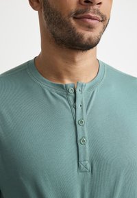 Camisa henley de algodón en un tono azul verdoso claro, con escote redondo, placket de tres botones y una superficie texturizada. Presenta detalles cosidos en el cuello.