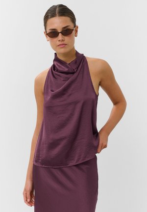 Femme portant un débardeur violet foncé à col bénitier et une jupe assortie avec des lunettes de soleil fines marron, debout devant un fond clair uni.