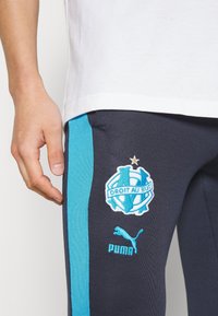 Námořnické sportovní legíny s světle modrými bočními panely. Obsahují vyšívané logo Marseille a značku Puma. Materiál vypadá elasticky.