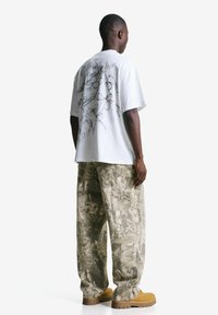 Man staat met zijn rug naar de kijker, gekleed in een witte T-shirt met abstracte print, camouflage-patroon wijde broek en tan kleurige werkschoenen tegen een witte achtergrond.