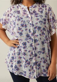 Kvinna klädd i en kortärmad, knappad vit blus med blå och rosa blommönster, med handen vilande på höften, tillsammans med mörka byxor.