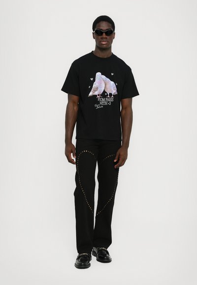 Camiseta negra con un gráfico colorido de paloma y texto. Pantalones negros con detalles de estrellas doradas a lo largo de las costuras. Zapatos negros y gafas de sol.