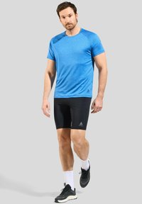 T-shirt de sport bleu à manches courtes, col rond et logo discret. Associé à un short noir ajusté et des baskets noires, avec des chaussettes blanches.