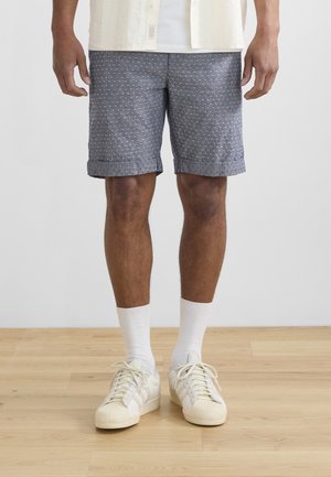 Mand iført blå mønstrede shorts, hvide crew-sokker og cremefarvede sneakers stående på trægulv mod en ensfarvet hvid væg.