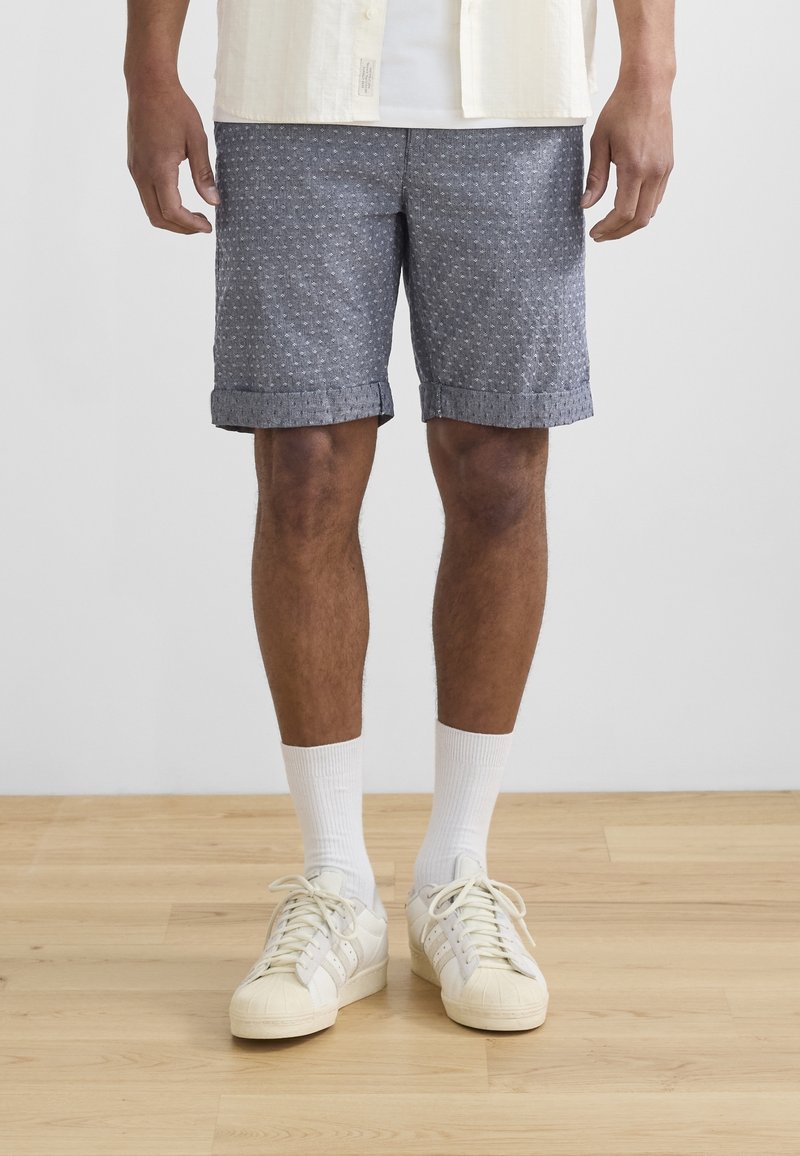 Homme portant un short bleu à motifs, des chaussettes blanches montantes et des baskets couleur crème, debout sur un sol en bois devant un mur blanc uni.