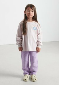 Svijetloružičasta košulja dugih rukava s plavim natpisom, uparen sa ljubičastim flis hlačama i bež klocama s ventilacijskim rupicama. Jednostavna, udobna kombinacija.