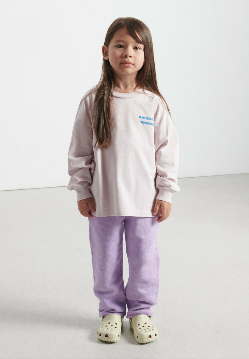 Svijetloružičasta košulja dugih rukava s plavim natpisom, uparen sa ljubičastim flis hlačama i bež klocama s ventilacijskim rupicama. Jednostavna, udobna kombinacija.