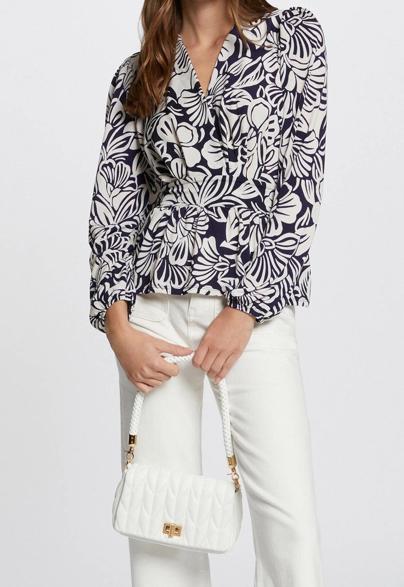 Femme portant un chemisier à fleurs bleu marine et blanc avec un pantalon blanc, tenant un sac à main matelassé blanc avec une bandoulière tressée et un fermoir doré.