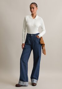 Witte geribde lange mouw top met een kraag, gecombineerd met donkerblauwe high-waisted wijde jeans met manchet en een bruine geblokte bandana.