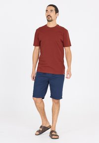 Herrklädsel med en bordeauxfärgad t-shirt med kort ärm, marinblå shorts med uppvikt fåll och bruna sandaler med spänningsdetalj.