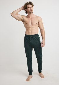 Donkergroene joggingsbroek met een trekkoord in de taille, slim fit en details van zijkanten. Geen zichtbare zakken. Model staat op blote voeten.