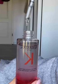 Duidelijke fles met Kérastase haarserum met een pipet; verloop van helder naar roze, reliëf logo en tekst, glad oppervlak.
