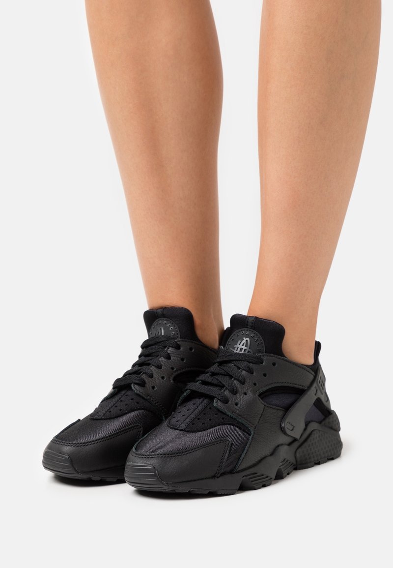 Jalat memakai sneakers atletik hitam dengan sol bertekstur dan tali sepatu, difoto di latar belakang putih polos.