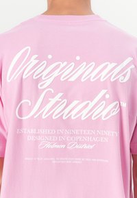 Lyserød bomulds t-shirt med stor hvid tekst på ryggen: "Originals Studio," med yderligere detaljer nedenfor i mindre skrifttype.