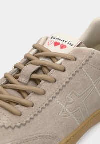 Μπεζ σουέτ αθλητικά παπούτσια με καφέ κορδόνια, έντονο ραφές και γλώσσα με επωνυμία που διαθέτει κόκκινες καρδιές. Σόλα από καουτσούκ.