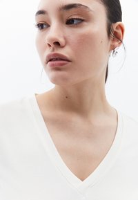 Top de color blanco roto con cuello en V y mangas cortas, confeccionado en una tela suave y ligera. La modelo lleva un arete de plata simple, mostrando un aspecto natural.
