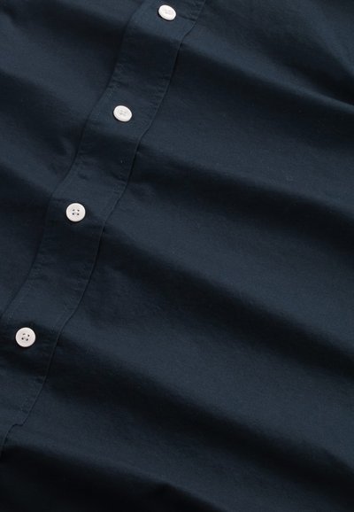 Tessuto in cotone blu navy con un leggero lucido, caratterizzato da una fila verticale di cinque bottoni in plastica bianca lungo il centro. Texture morbida e liscia.