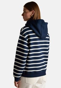 Žena v námornícky modrom a bielom pruhovanom mikine s kapucňou stiahnutou dole, v kombinácii s svetlými nohavicami, stojí smerom doľava na jednolatom pozadí.