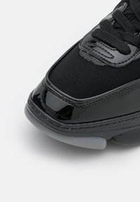 Gros plan sur l'avant d'une basket noire avec un cuir verni brillant, une tige en tissu, des lacets noirs et une semelle texturée sur fond blanc.