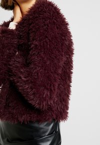 Burgundská umělá kožušina s nadýchanou texturou, s krátkým střihem. V páru s hladkou černou koženou sukní, zdůrazňující kontrast v materiálech.