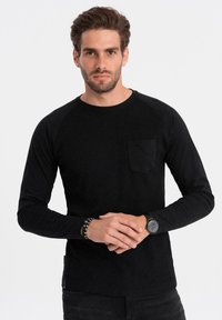 Svart långärmad ribbad tröja med rund halsringning, med en bröstficka och slät struktur. Modellen bär den med mörka byxor.