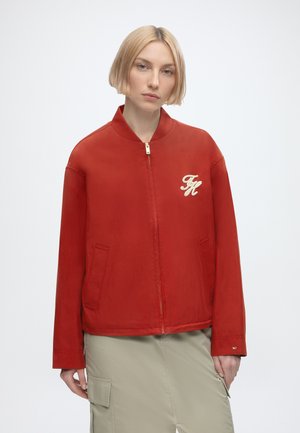Jonge vrouw met kort blond haar, draag een rode jas met ritssluiting met een crème "FH" logo op de borst en lichtbeige cargobroek.