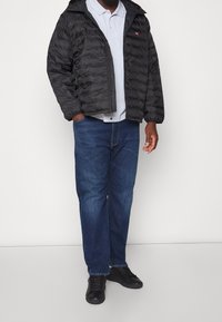 Man i svart pufferjacka, vit polo-skjorta, blå jeans och svarta sneakers står framför en ljusbakgrund.