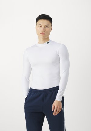 Top bianco a maniche lunghe realizzato in tessuto morbido con collo alto, abbinato a pantaloni della tuta blu scuro con strisce laterali e un logo.
