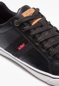 Musta lenkkikenkä, jossa on teksturoitua nahkaa ja kangasdetailit. Varustettu ruskealla nahkatarralla, mustilla nauhoilla ja valkoisella kumipohjalla, jossa musta reunus.