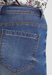 Denimfarkut keskisinisellä värillä, joissa on takatasku ja kontrastinen oranssi ompelu. Kangas on sileäpintainen.