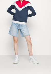 Felpa con zip color block blu navy e rossa con dettagli bianchi, abbinata a pantaloni shorts in denim blu chiaro con orlo sfrangiato. Sneakers bianche completano il look.