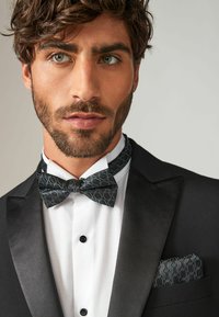 Tuxedo with black satin lapels, white textured shirt, and patterned black bow tie. Pocket square matches bow tie design.