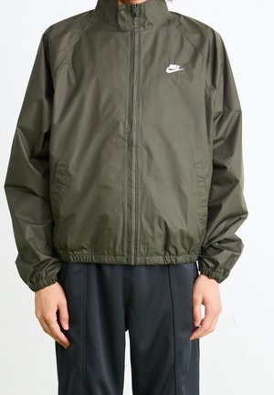 Homme portant une veste zippée Nike vert olive avec des poignets élastiques et un pantalon noir, se tenant devant un fond blanc.