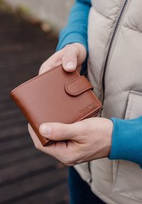 Brown leather wallet with a smooth texture, featuring a snap closure and embossed "VUCH" logo, held by hands in a casual setting.