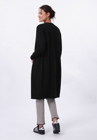 Black long cardigan with long sleeves and pockets, made of a textured fabric; paired with gray jeans and black-and-white sneakers.
