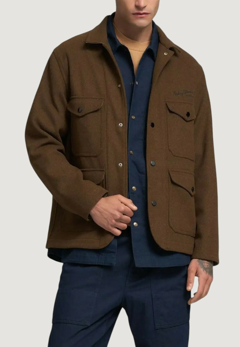 Giacca in misto lana marrone con quattro tasche frontali, bottoni a pressione e design casual, indossata sopra una camicia blu.