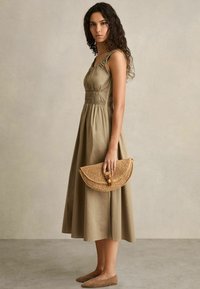 Khaki katoenen jurk met gerimpelde details, gecombineerd met een geweven clutch en taupe platte schoenen. Heeft een aansluitend bovenlijfje en een vloeiende rok.