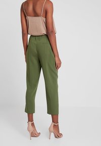 Pantalones verdes a medida con corte recto, cintura alta, dos bolsillos traseros y costuras sutiles, combinados con sandalias de tacón desnudo.