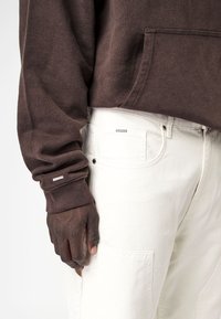 Una mano de piel oscura descansa junto al bolsillo de unos pantalones blancos, vistiendo una camiseta de manga larga marrón con un bolsillo frontal y una pequeña etiqueta de la marca en el puño.