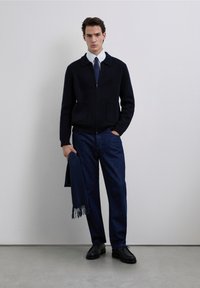 Mariene ritsvest met een kraag, gecombineerd met donkere denimbroek en zwarte schoenen. Een bijpassende marineblauwe sjaal hangt over de arm.