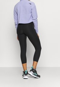 Osoba w czarnych spodniach 3/4, lawendowej polarowej kurtce z logo The North Face i czarno-błękitnych butach sportowych, stojąca bokiem.